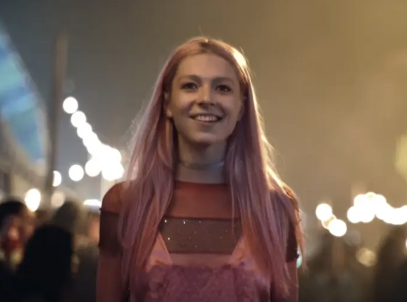 Hunter Schafer smiles, wearing a stylish sheer top and pink outfit at an outdoor event with string lights in the background