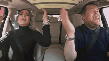 Jennifer Lopez and James Corden enthusiastically sing together in a car with their hands raised during a Carpool Karaoke segment