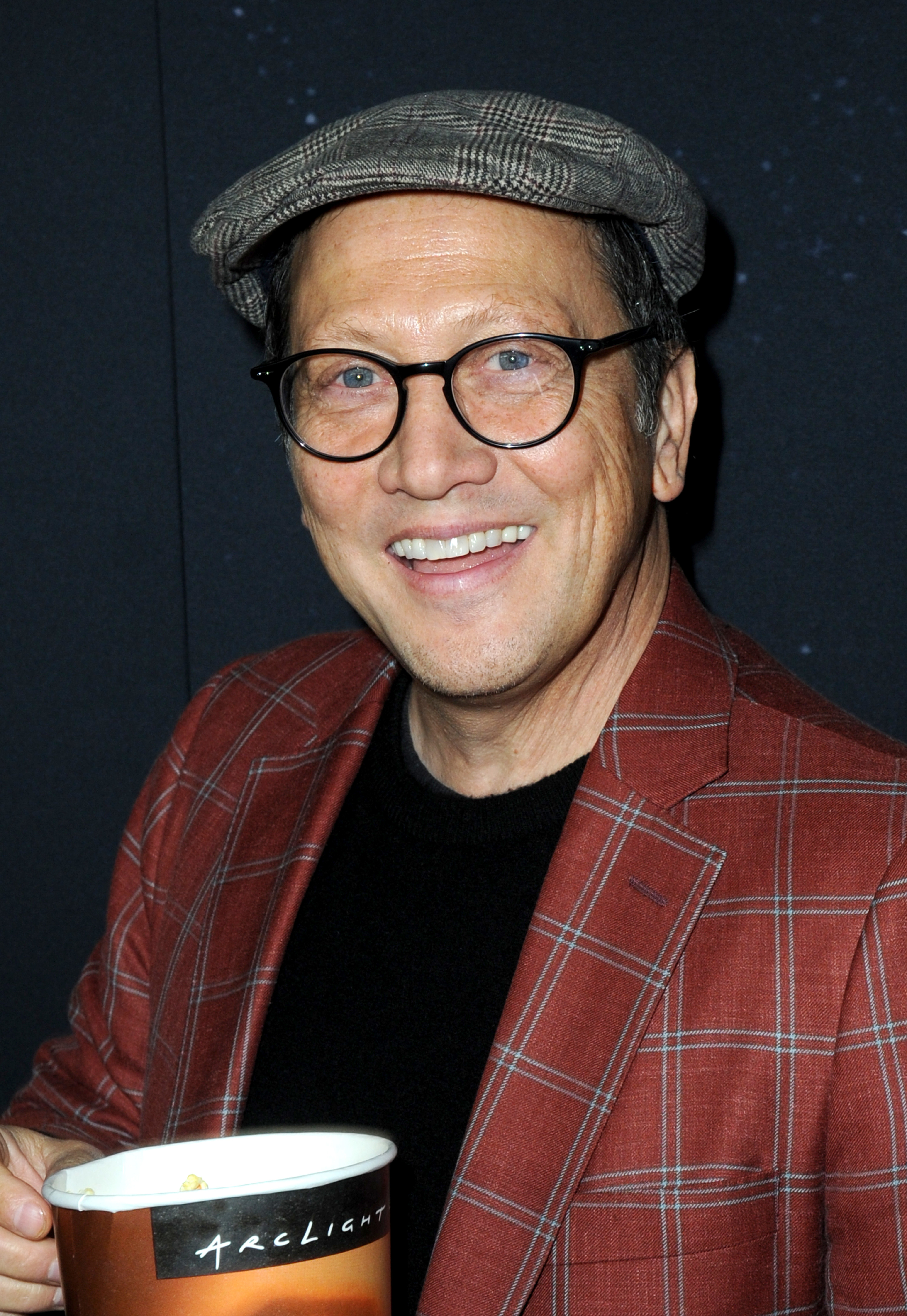 Rob Schneider smiles while holding a cup, wearing a plaid blazer, black shirt, round glasses, and a flat cap