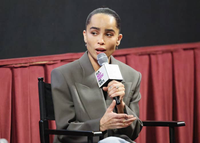 Zoe Kravitz speaks into a microphone while sitting on a stage, wearing a blazer, with a serious expression