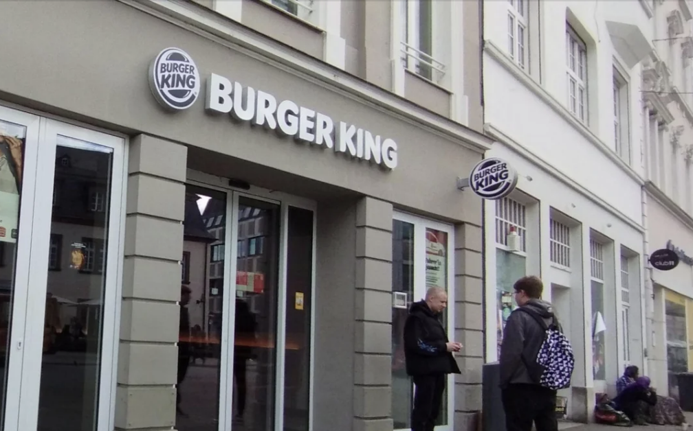 Dua orang berinteraksi di luar restoran Burger King dengan logo yang terlihat. Satu orang duduk di dekat gedung