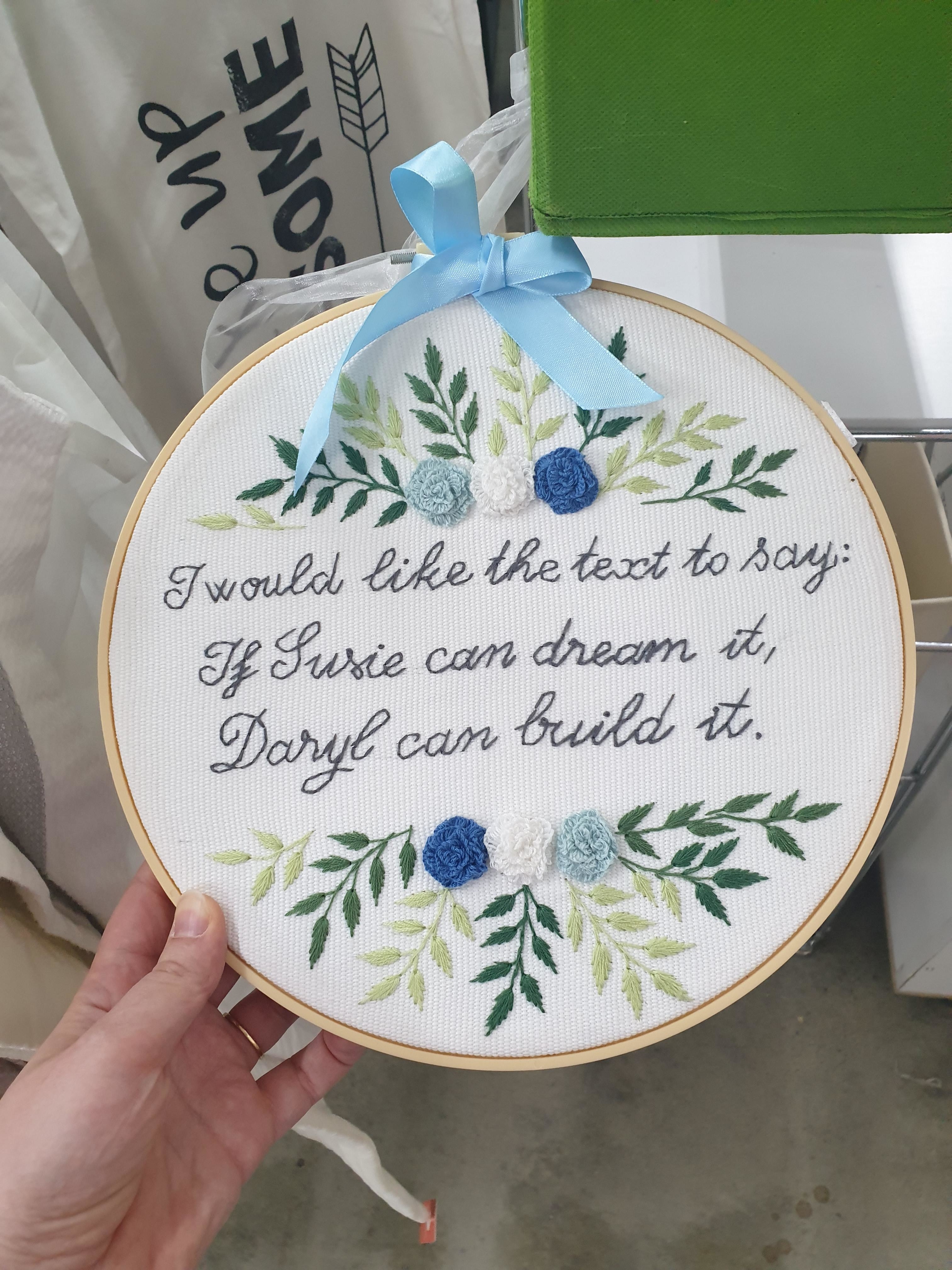 A hand holds an embroidery hoop featuring floral designs and the text: "If Susie can dream it, Daryl can build it."