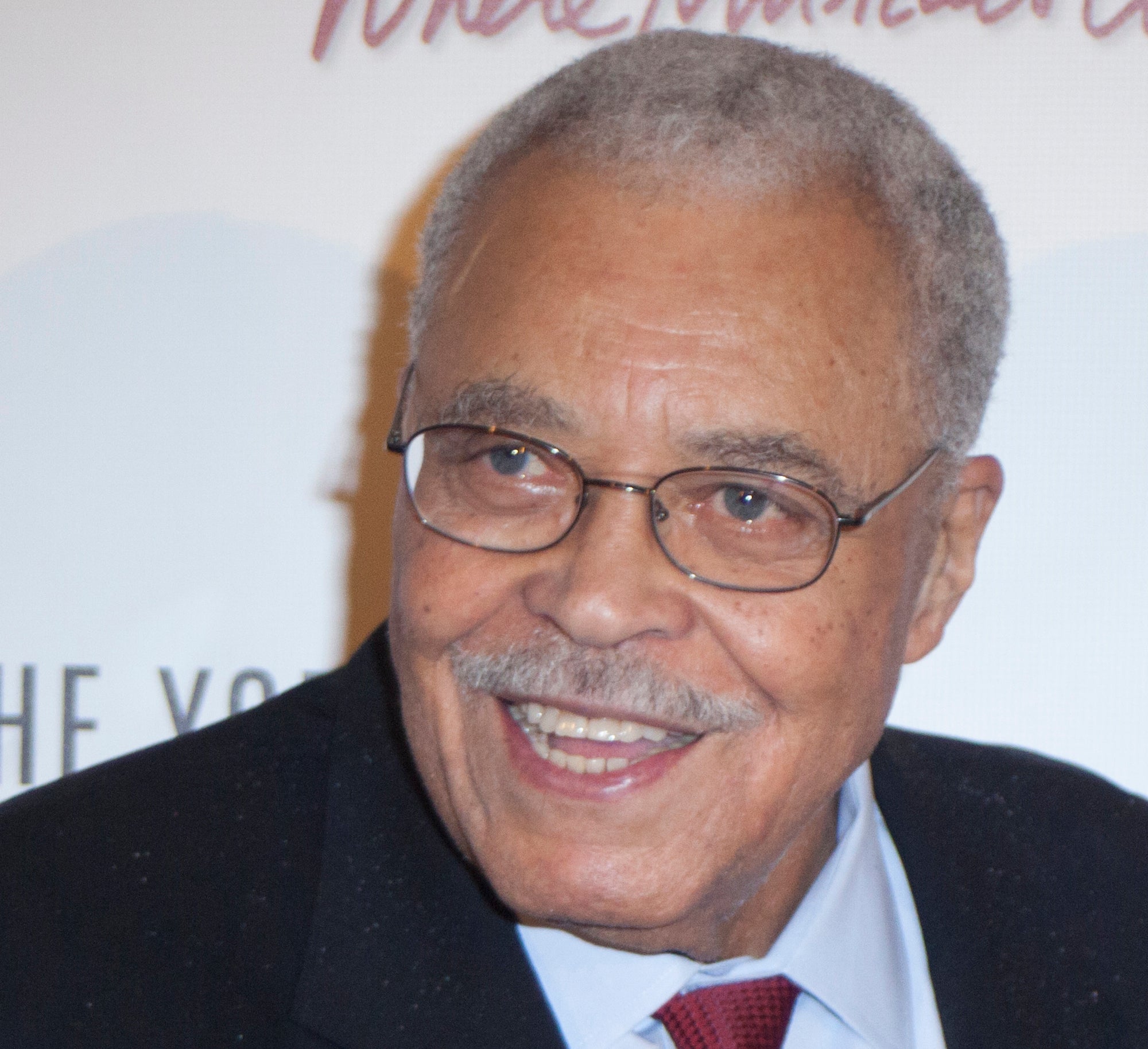 James Earl Jones smiles while wearing a black suit, light blue shirt, and red tie at a public event backdrop with "Where Musicals…" printed