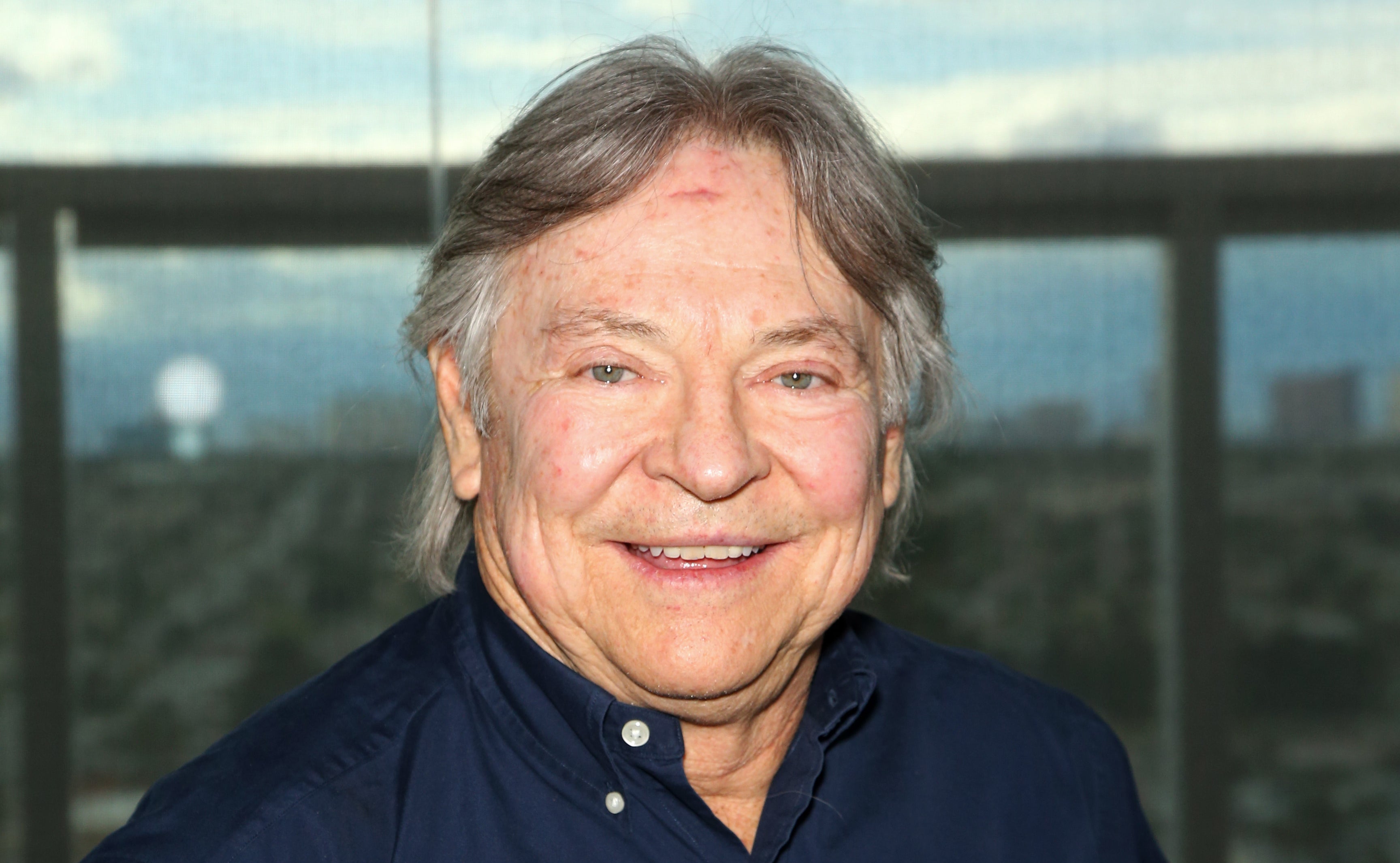 A man, Mike Lookinland, smiles while standing indoors near a large window with an urban background. He is wearing a button-down shirt