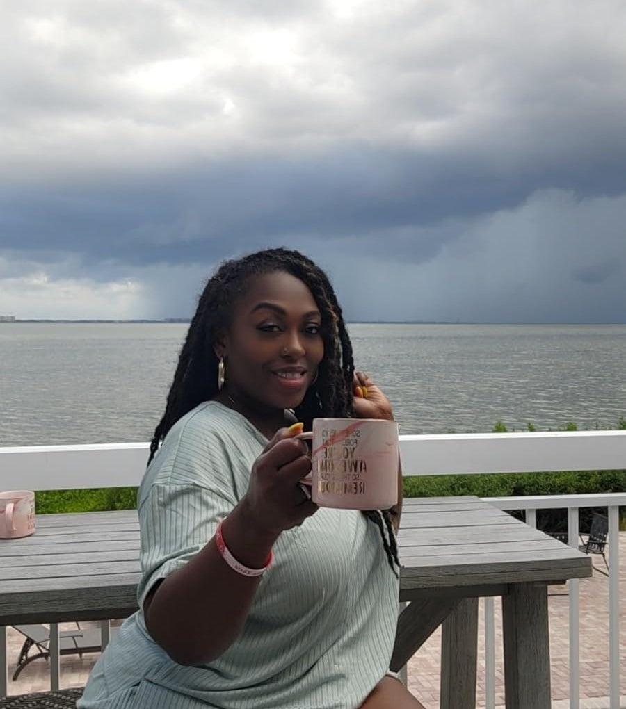 Person enjoying a drink on an outdoor terrace near the sea, holding a mug in one hand and smiling at the camera, with a cloudy sky and ocean in the background, wearing the teal matching set