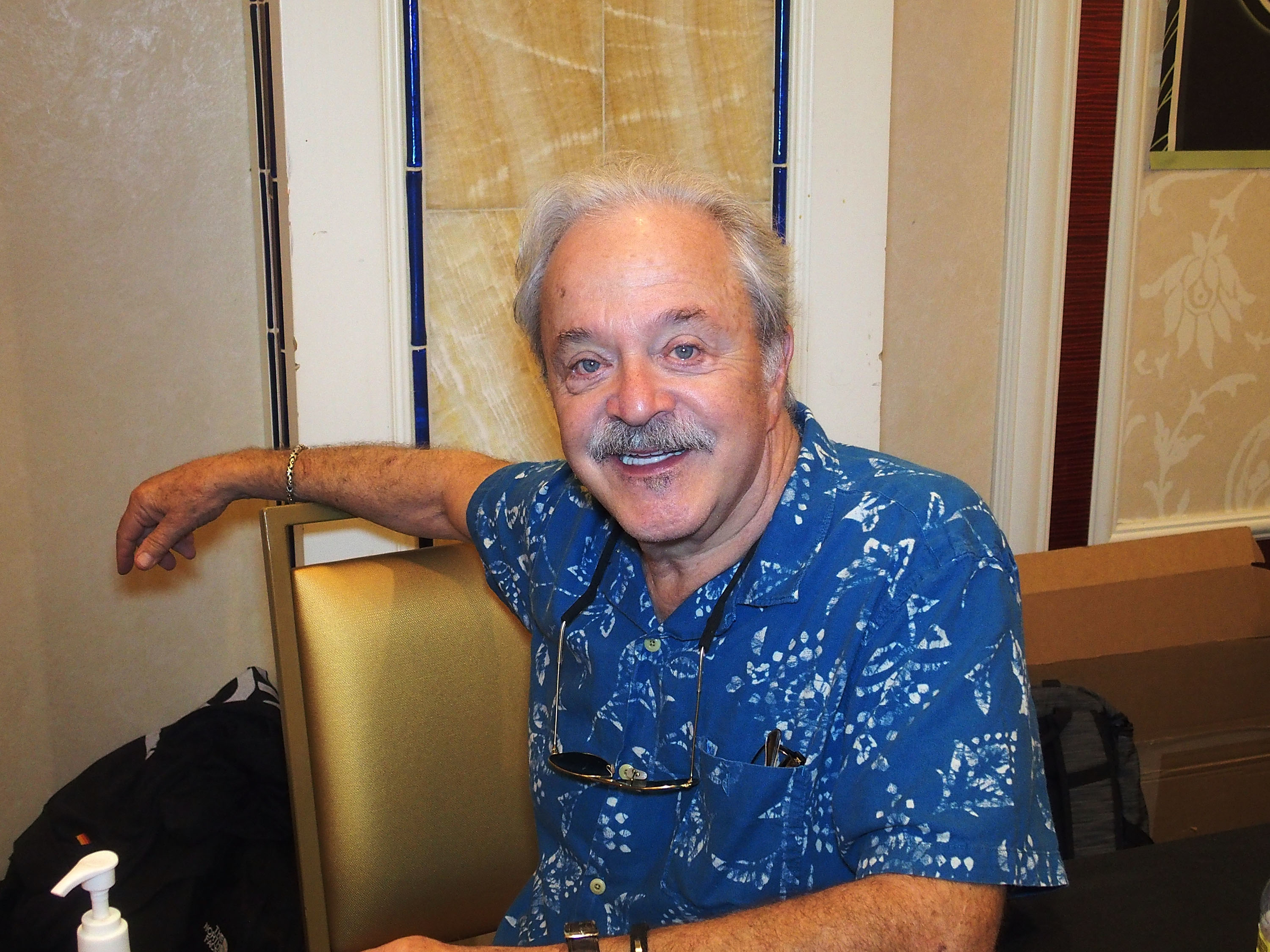 Jim Cummings, wearing a patterned short-sleeve shirt, sits at a table with an arm casually resting on the back of a chair, smiling broadly