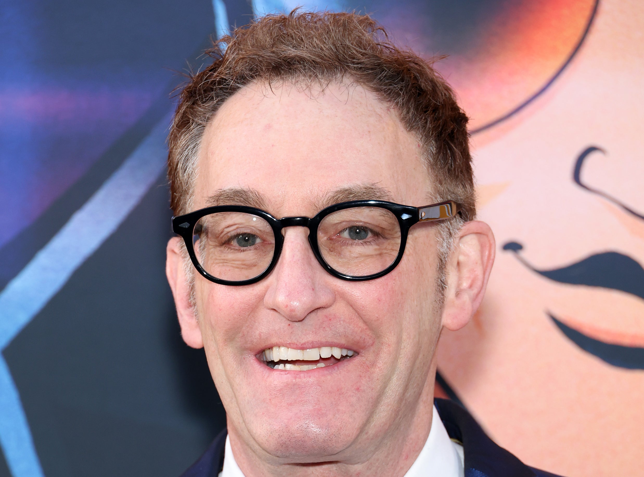 Tom Kenny wears a blue suit with a white shirt and a patterned tie, smiling on the red carpet at a TV and Movies event