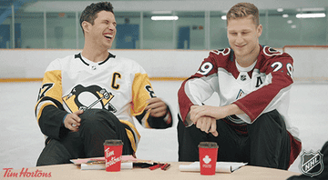 Sidney Crosby and Nathan MacKinnon sit at a table in an ice rink, laughing and drinking coffee, wearing their respective Pittsburgh Penguins and Colorado Avalanche jerseys