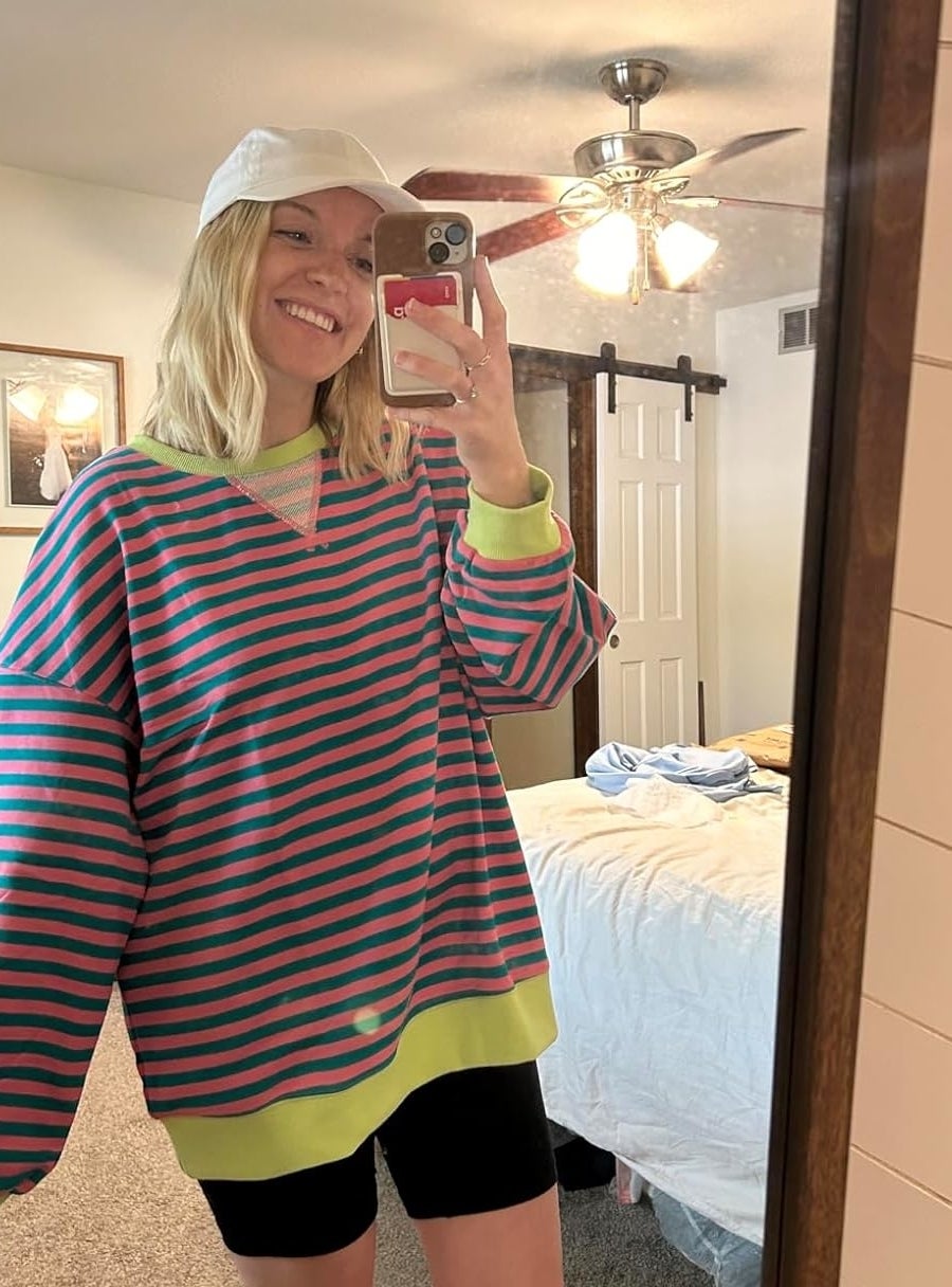 Reviewer in a striped oversized sweater, black shorts, and a white cap takes a mirror selfie in a bedroom with a ceiling fan and a bed in the background