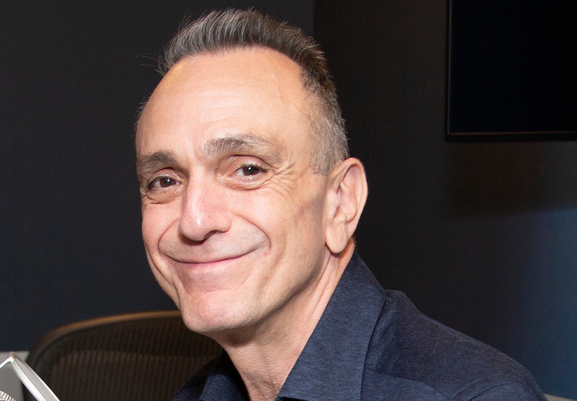 Hank Azaria smiling while seated in a chair, wearing a button-up shirt with sunglasses hanging from the neckline