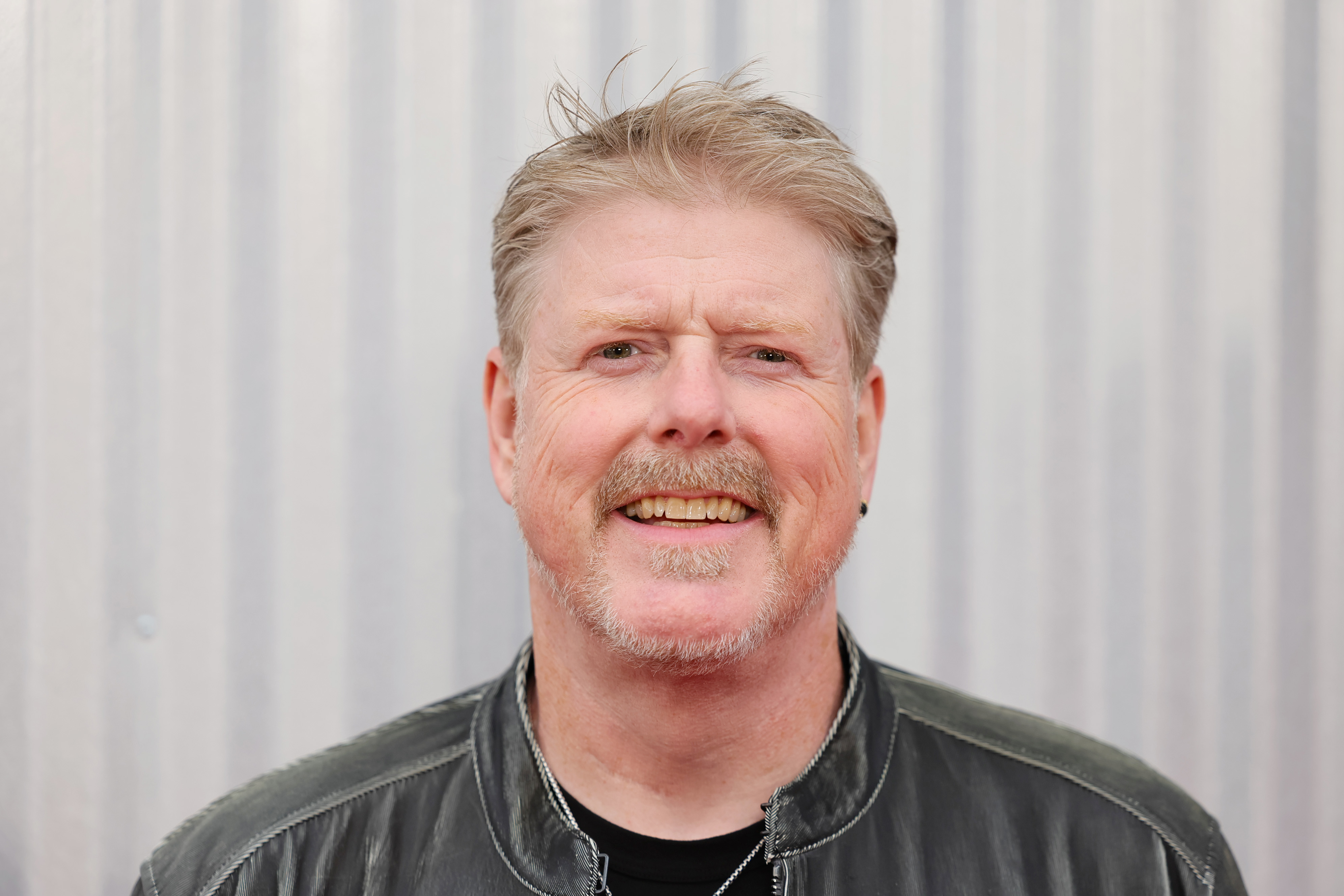 A man is smiling directly at the camera. He has short, light-colored hair and a mustache, and is wearing a leather jacket