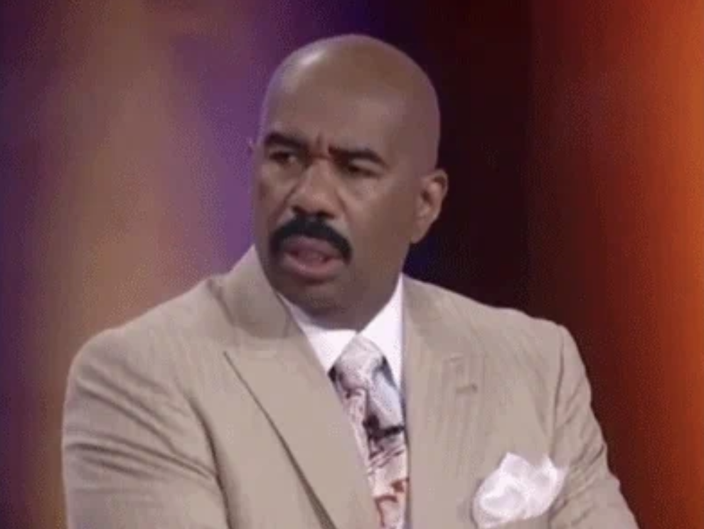 Steve Harvey dressed in a beige suit, white shirt, and patterned tie, looking to the right with a slightly surprised expression