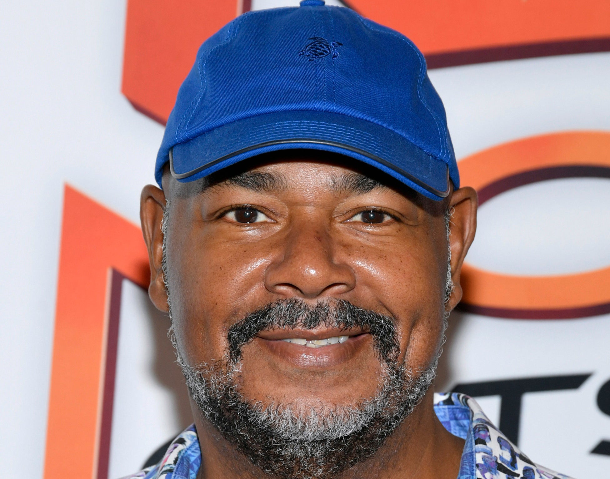 Kevin Michael Richardson wearing a blue baseball cap and a multicolored, patterned shirt smiles for the camera