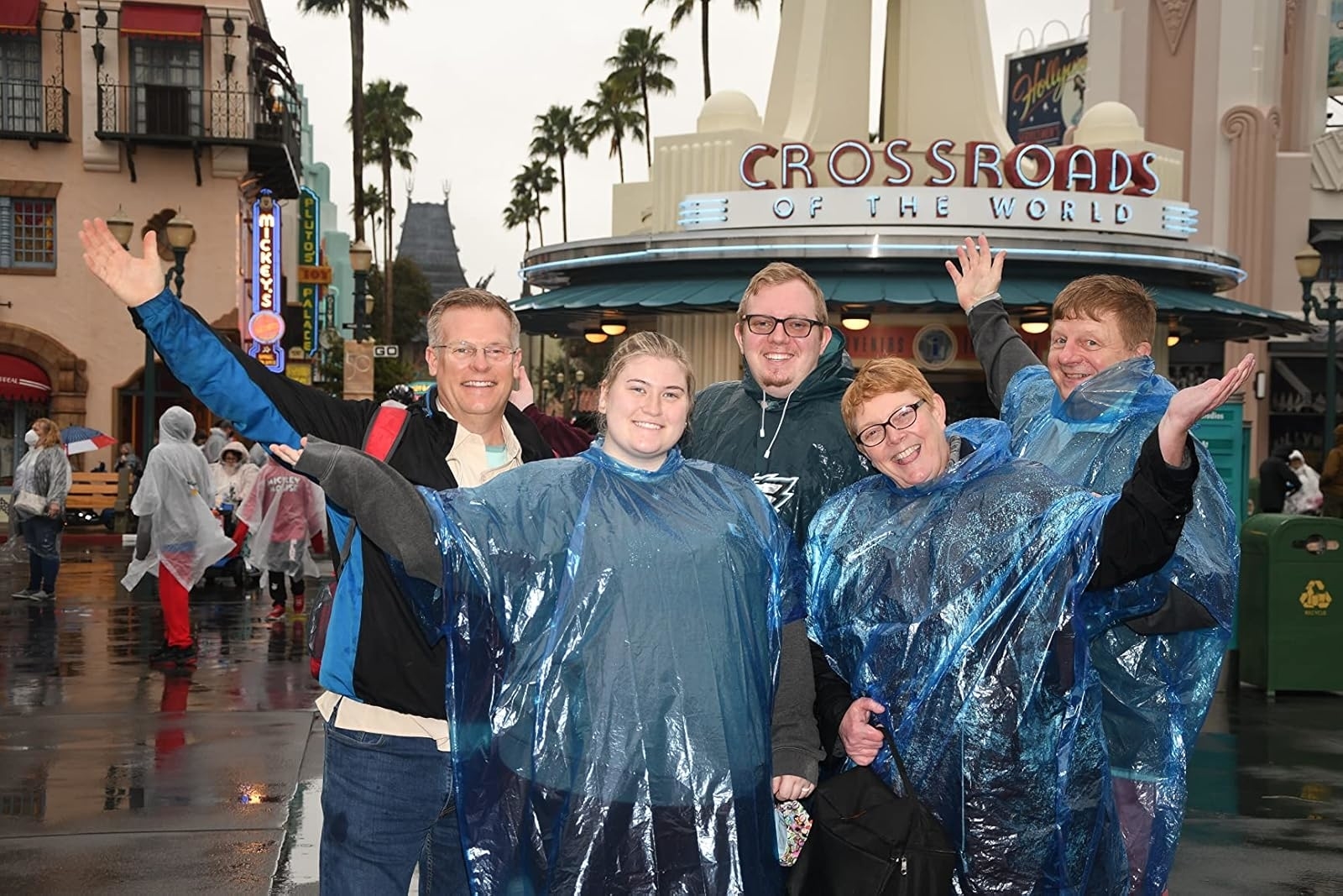 Five people in blue ponchos smile and raise their arms in front of the "Crossroads of the World" sign at an outdoor shopping area