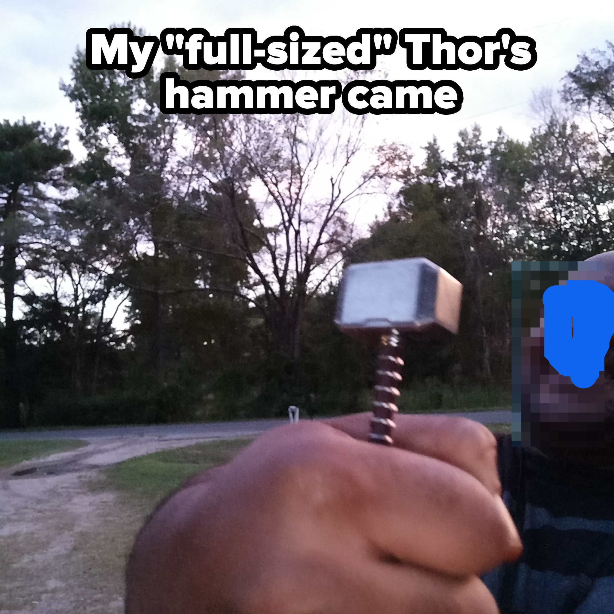 A smiling man holds a small replica of Thor's hammer Mjolnir in an outdoor setting with trees in the background