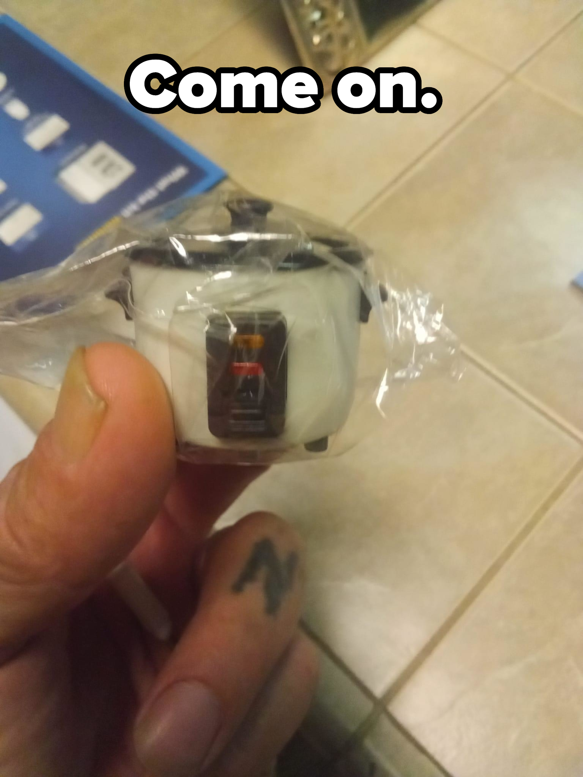 Person holding a miniature toy rice cooker still in its packaging. There is a tattoo on their finger