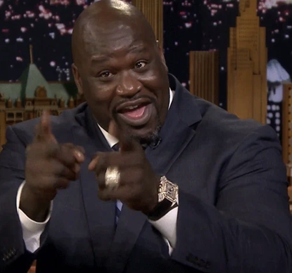 Shaquille O'Neal in a suit, smiling and pointing playfully towards the camera with both hands on a talk show set