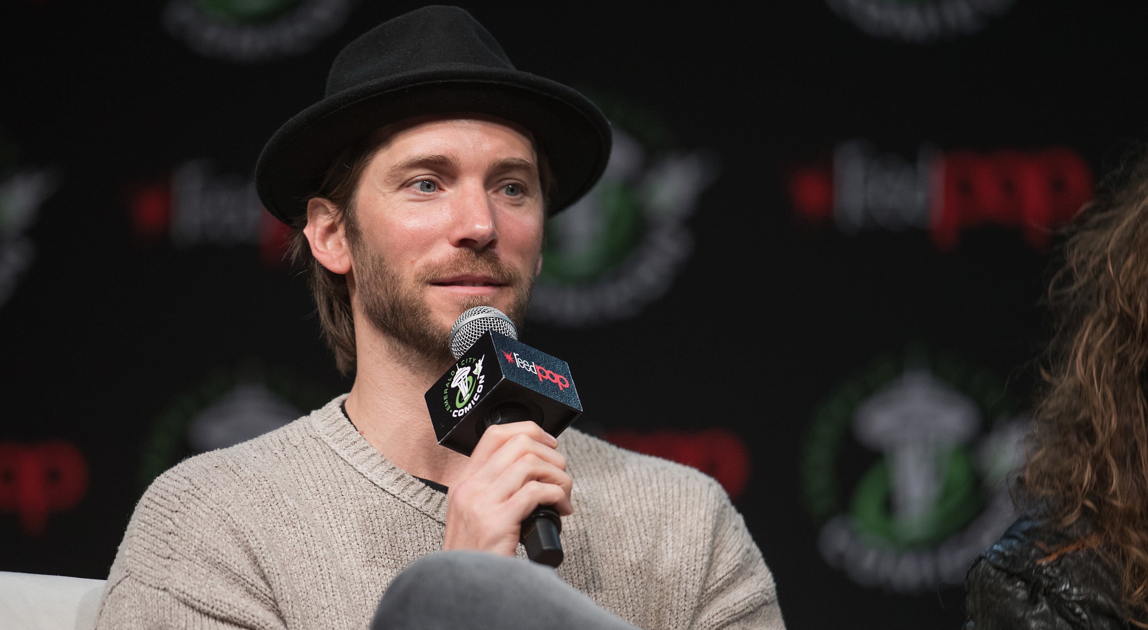 Troy Baker speaks into a microphone while sitting at a panel event. He is wearing a hat and a knit sweater