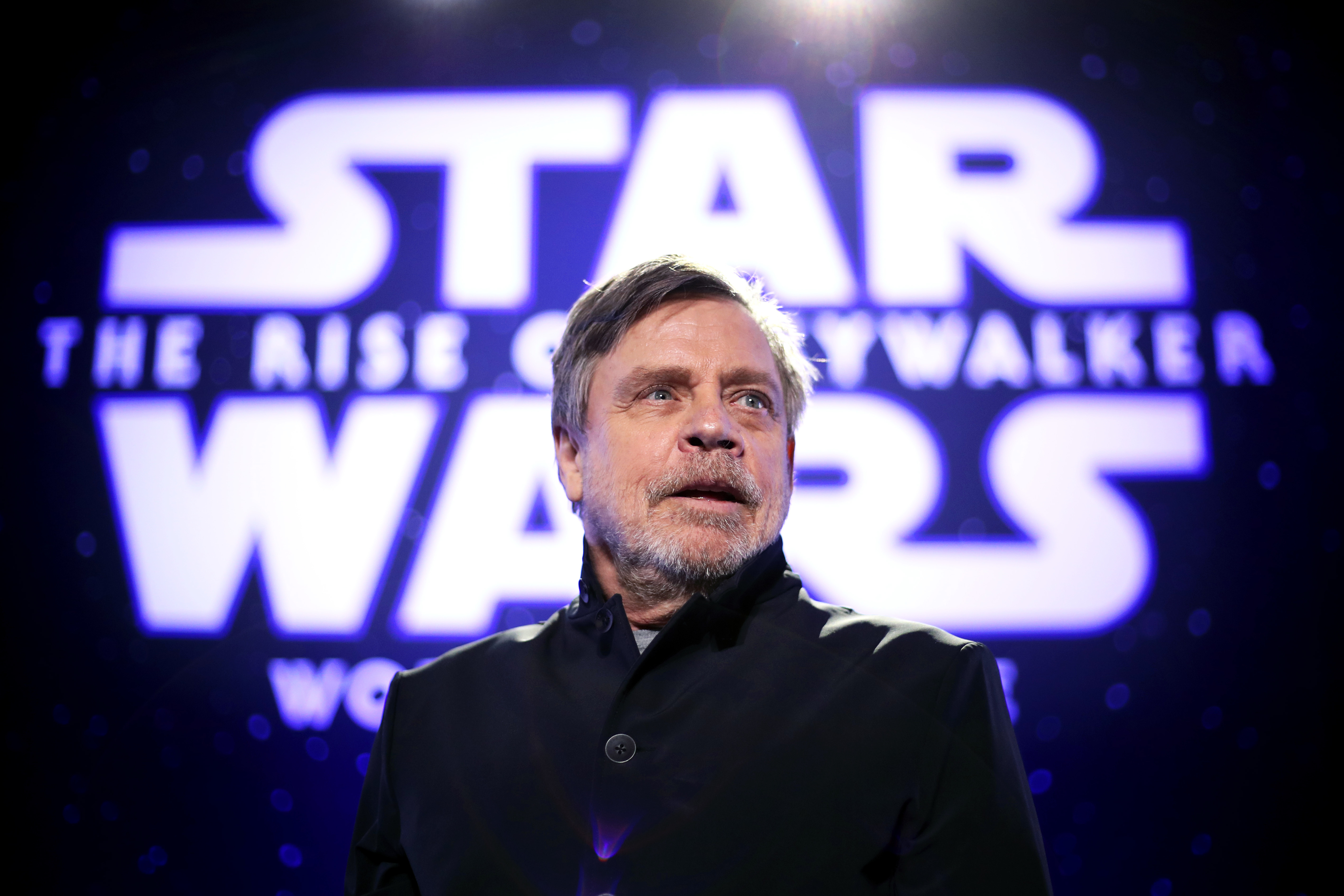 Mark Hamill stands in front of a "Star Wars: The Rise of Skywalker" poster wearing a dark jacket at an event