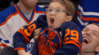 A child wearing an Edmonton Oilers jersey appears excited at a hockey game while being held up by an adult in the crowd