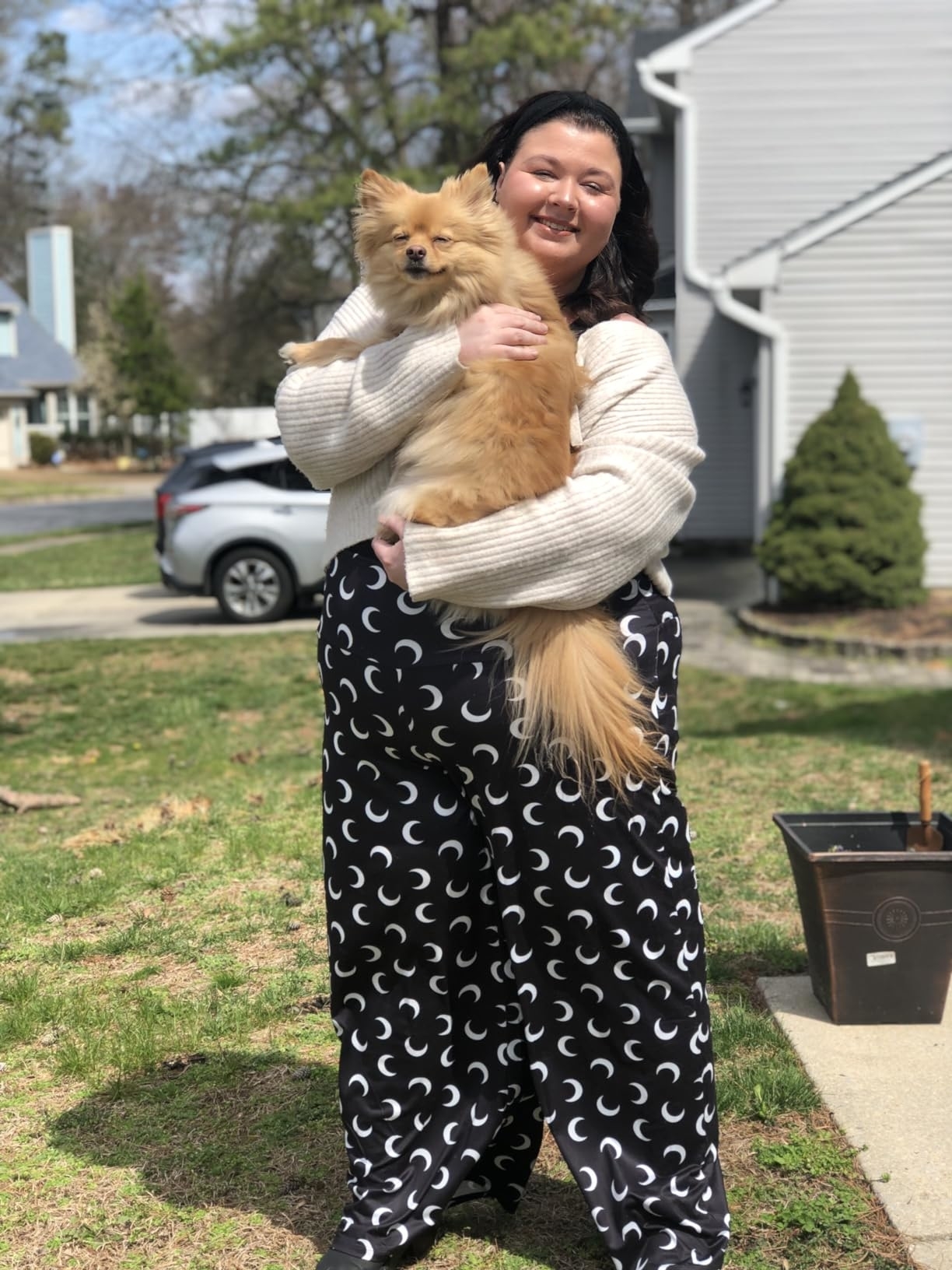 A smiling reviewer wearing patterned pants and a cozy sweater holds a fluffy dog outside in a residential area