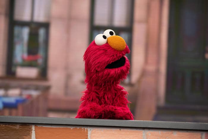 Elmo stands behind a brick wall, looking upwards with an open mouth