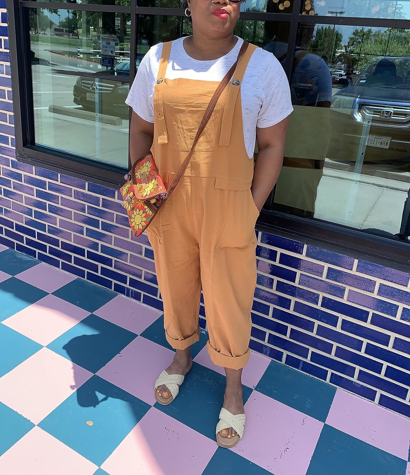 A person is seen standing on a checkered floor, wearing a white t-shirt, tan overalls, and woven sandals. They carry a floral crossbody bag