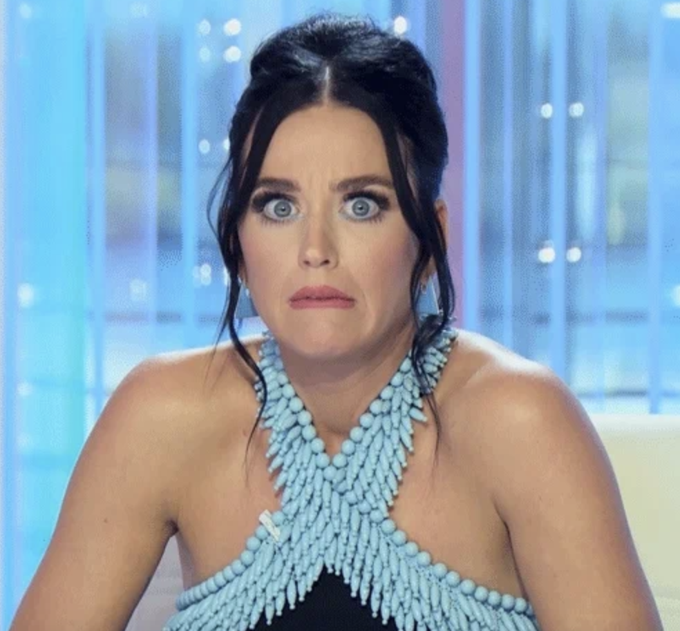 Katy Perry on a show set, making a surprised face, wearing a beaded halter top dress