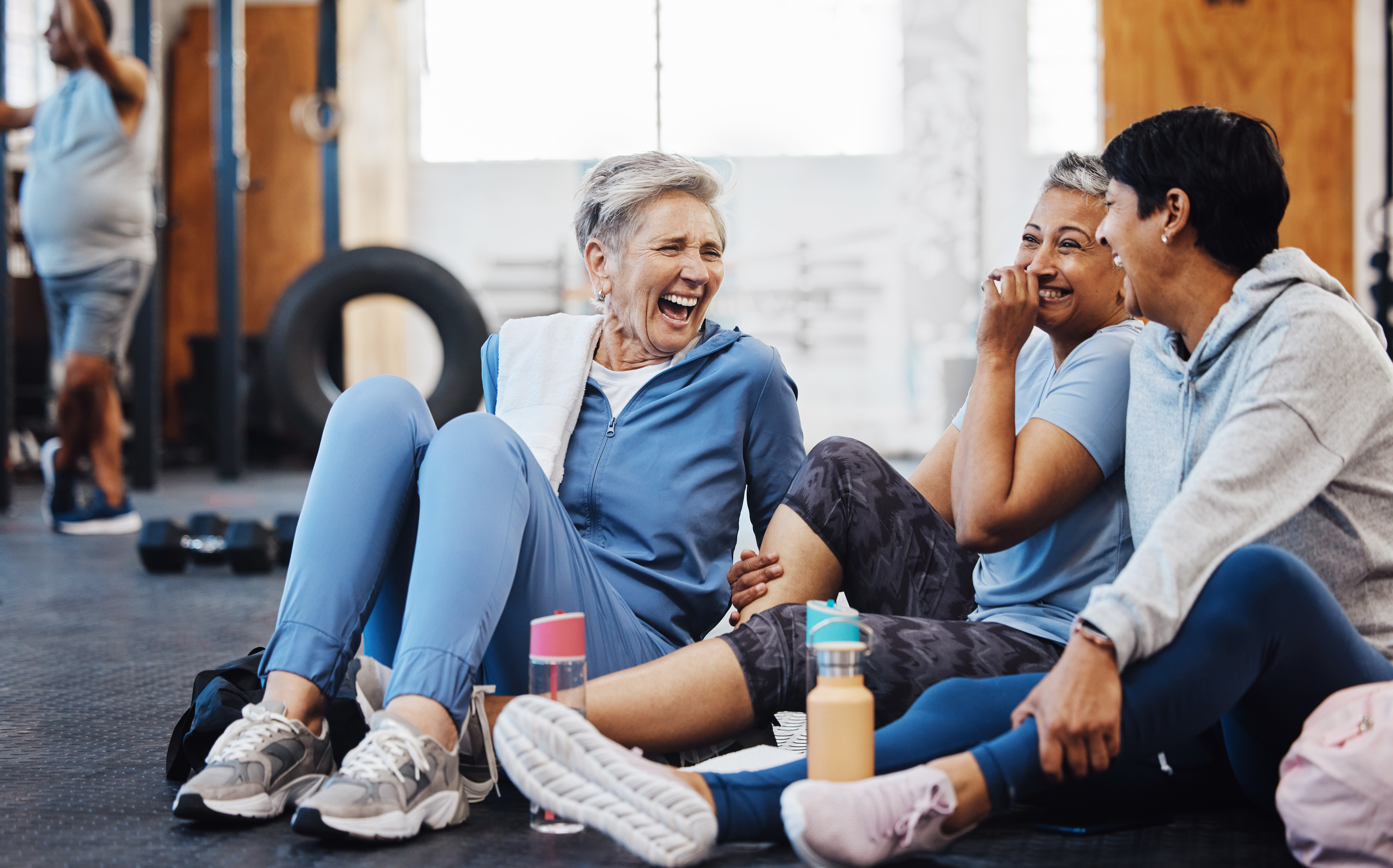 Women laughing and sitting on the floor in a gym, dressed in casual workout clothes, with water bottles around them