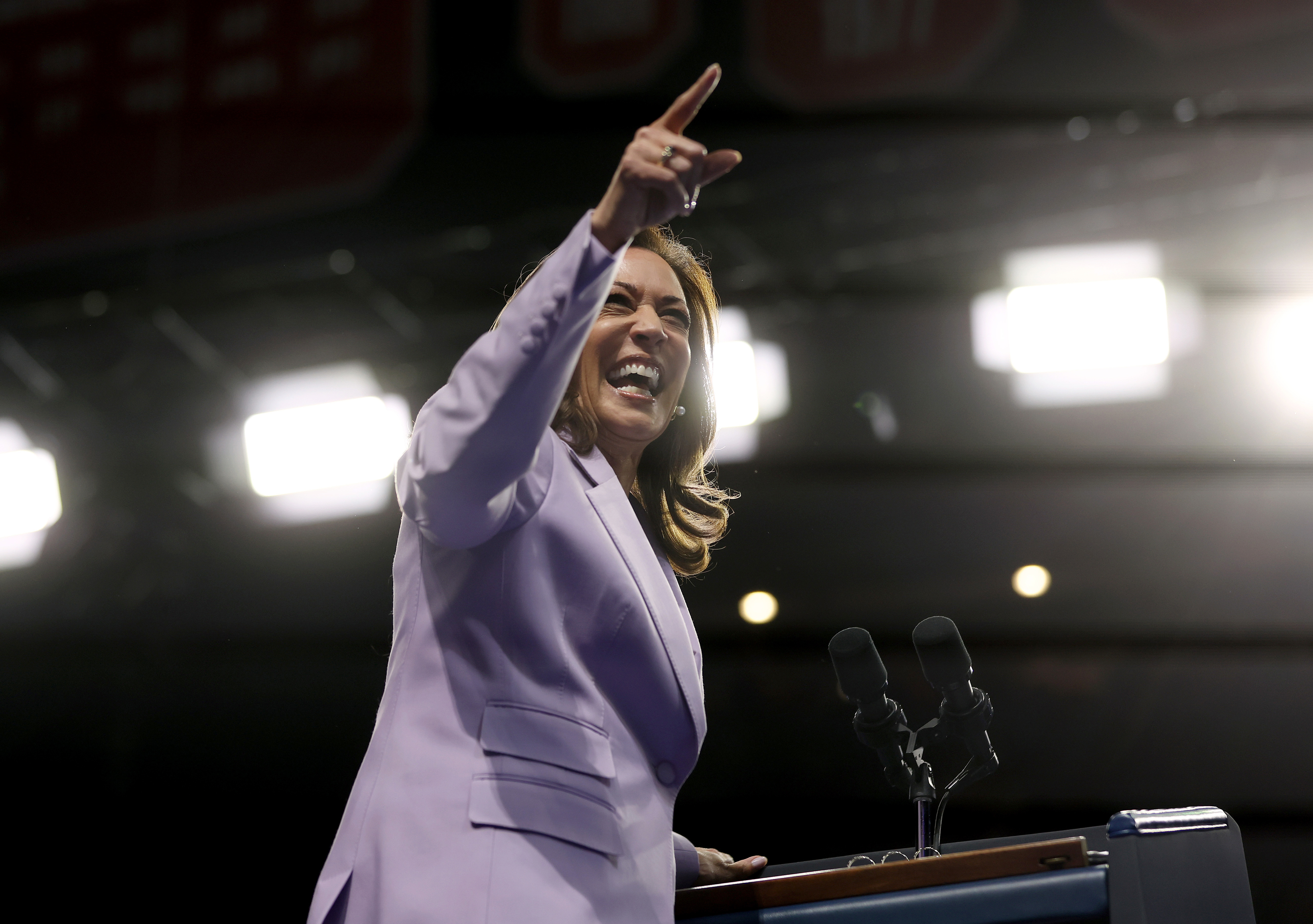 Kamala Harris wearing a light-colored suit, smiling and pointing upwards while standing behind a podium with microphones