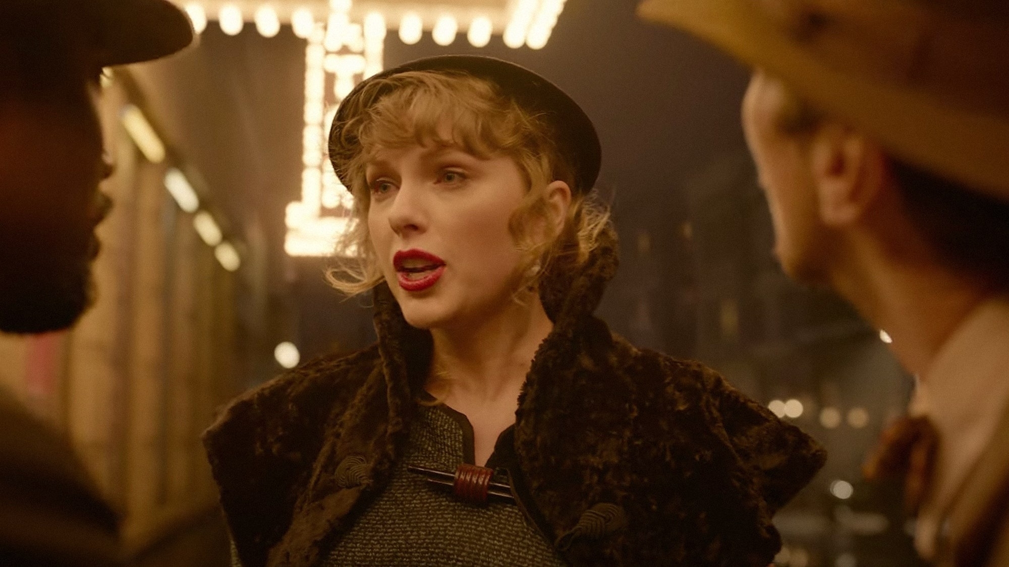 Taylor Swift in a vintage-style coat and hat speaking with two unidentified people in an urban night scene