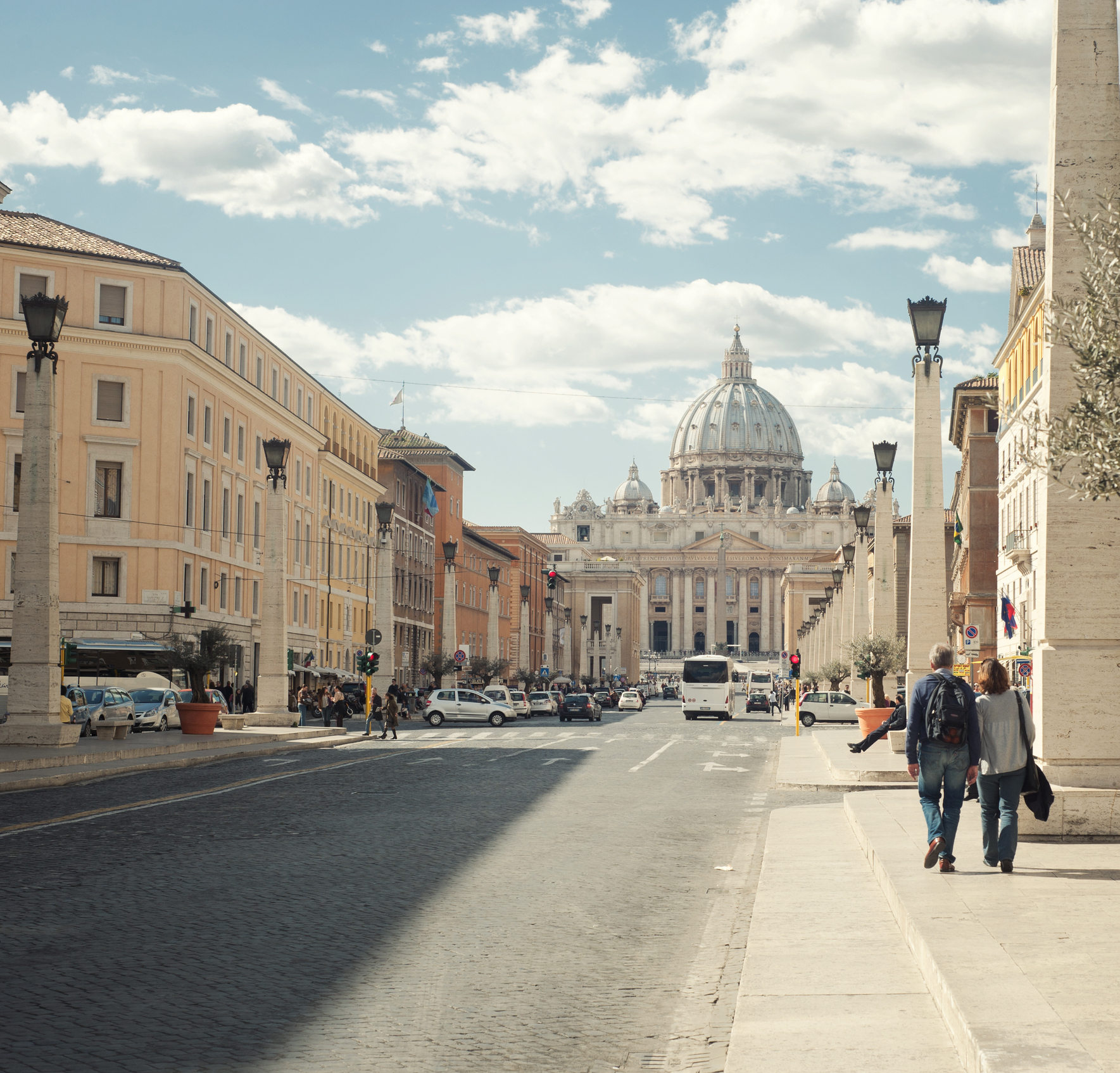Street view of Vatican City, showing St. Peter's Basilica in the background, with people walking on the sidewalk and cars driving on the road