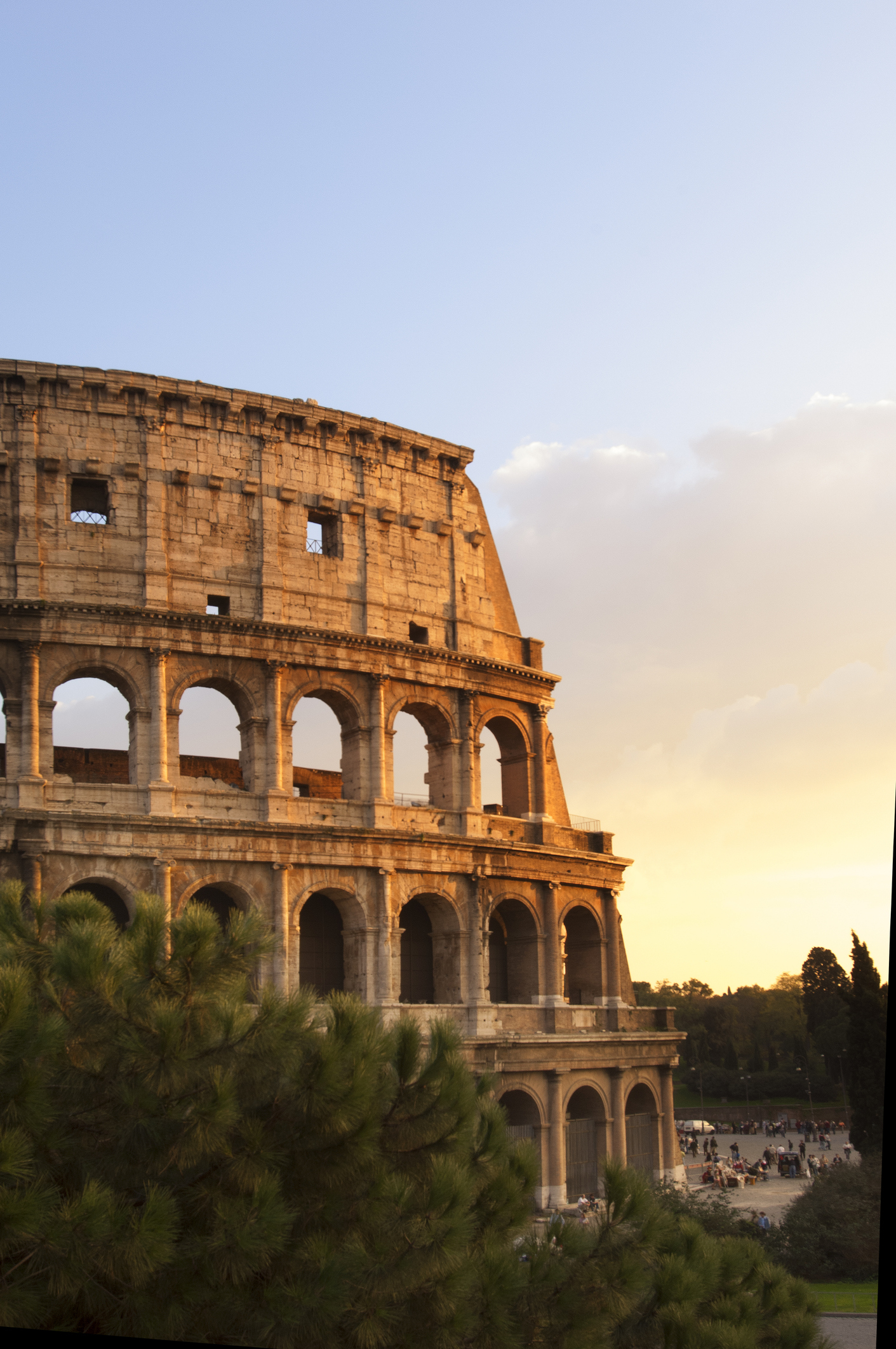 The image shows the Roman Colosseum at sunset, with the ancient amphitheater partially visible. A crowd can be seen gathering near the base. Trees are in the foreground