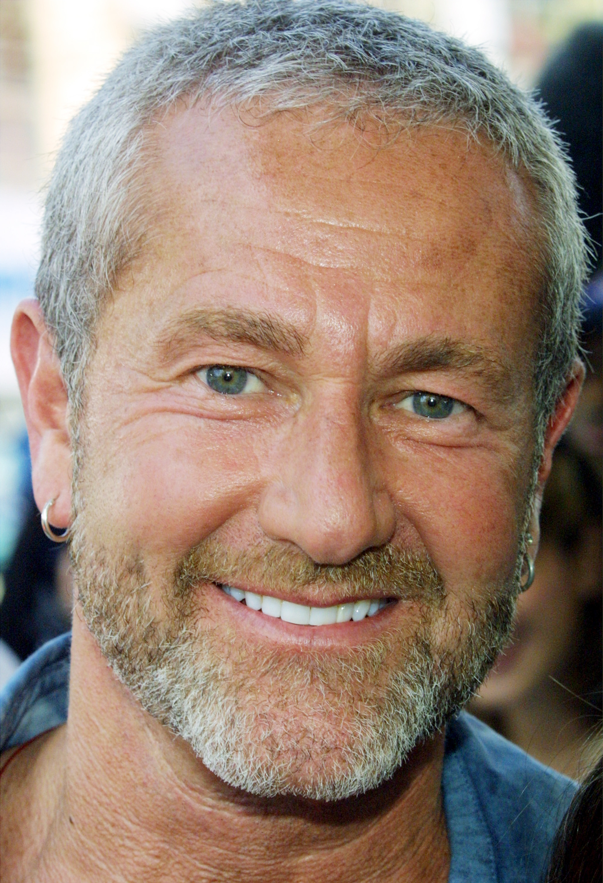 A man with short gray hair, a beard, and blue eyes smiles warmly at the camera. He is wearing an earring and a casual shirt