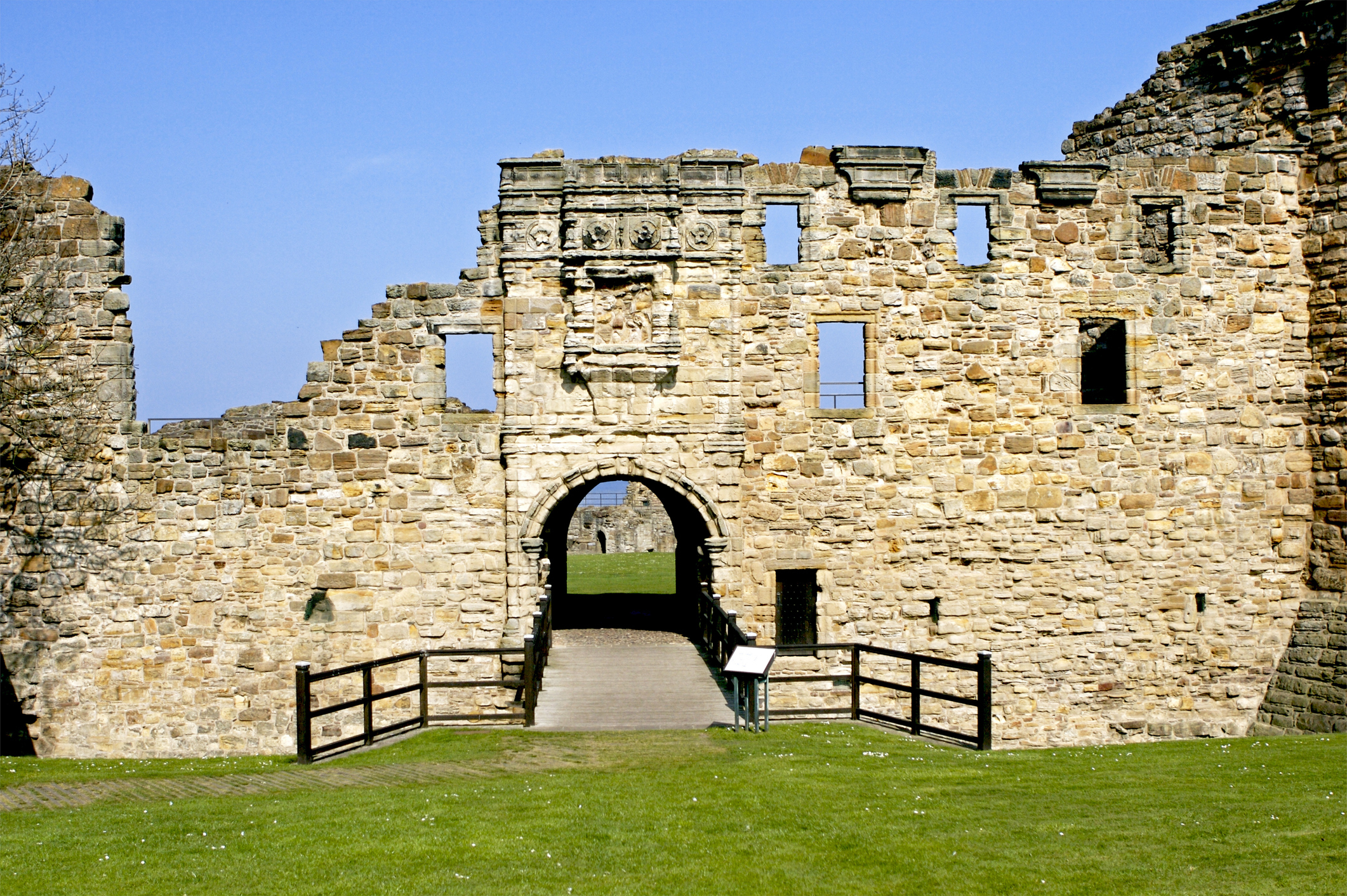 Ancient stone ruins with a central archway, surrounded by a grassy area and wooden fences. Bright sky in the background