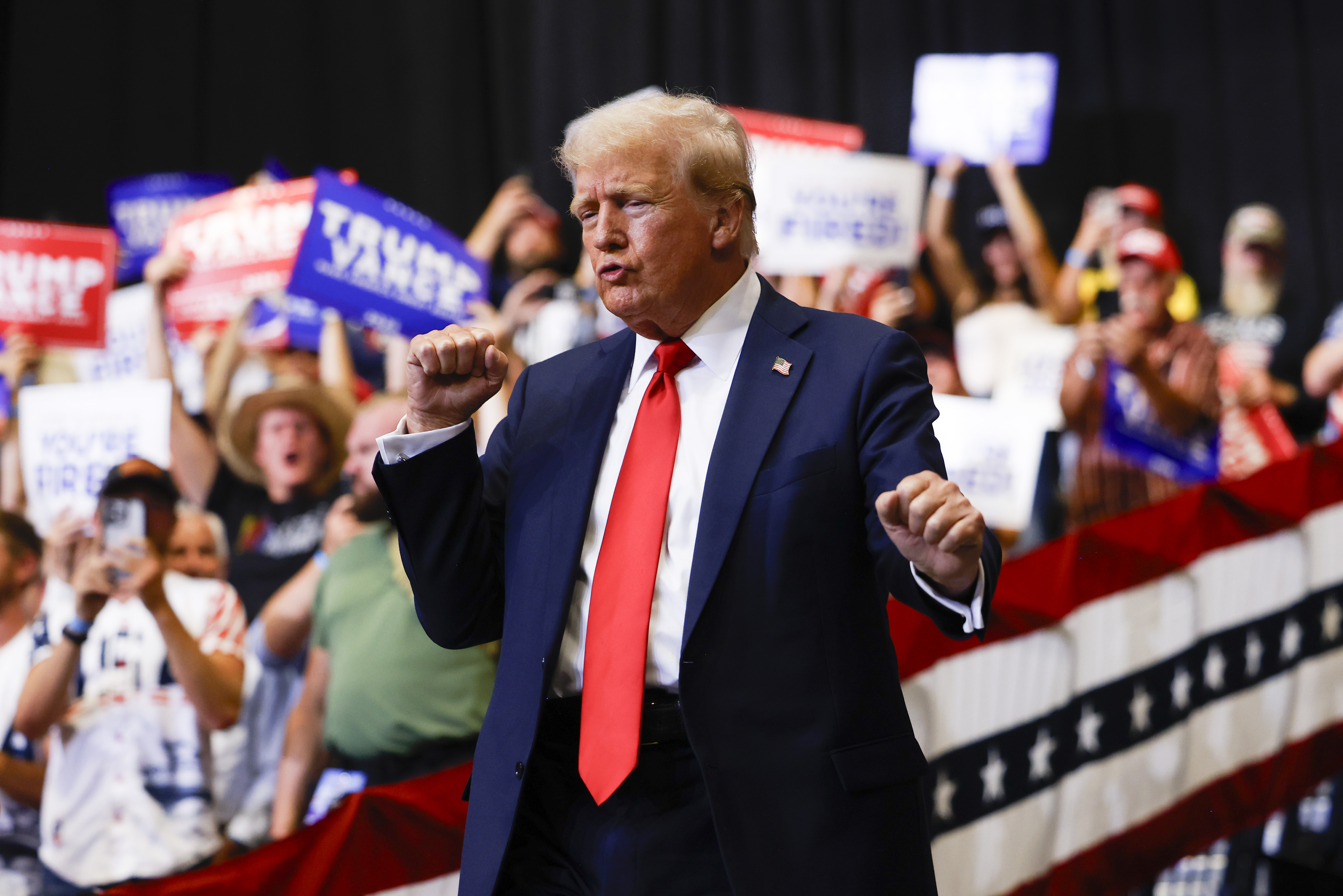 Donald Trump gestures with fists raised at a rally, surrounded by supporters holding signs
