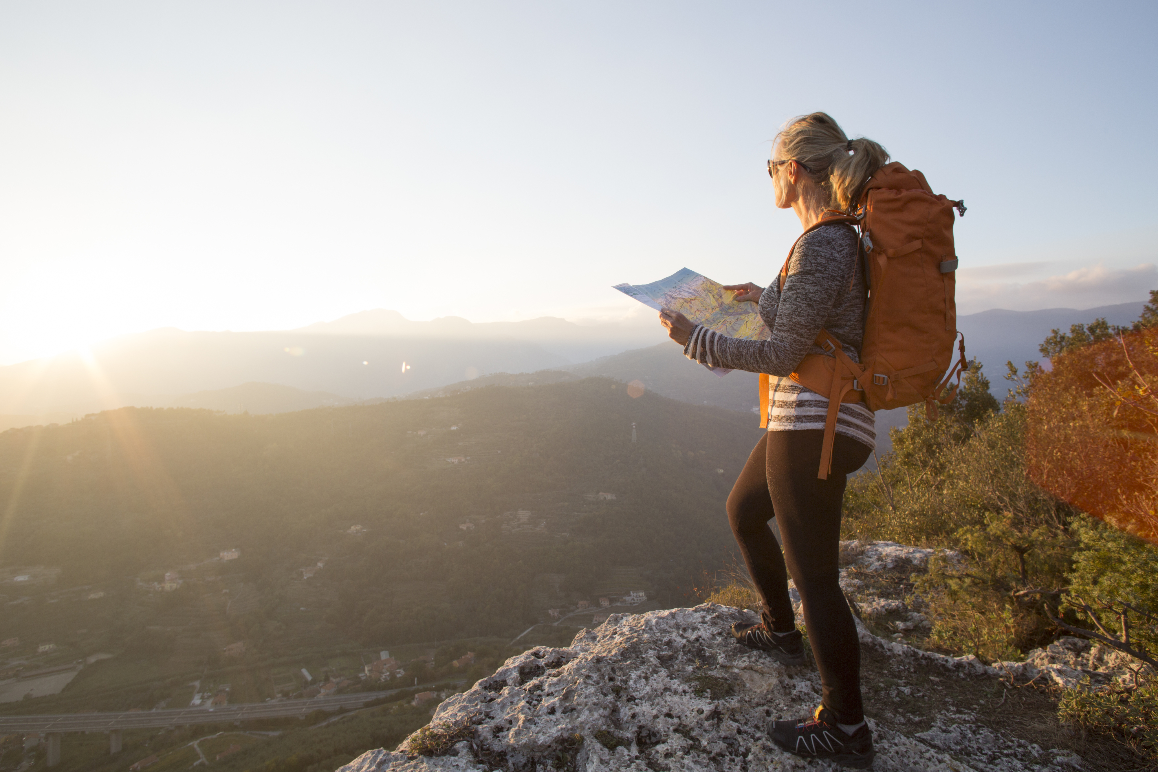 A woman stands on a mountaintop, wearing a backpack and looking at a map. The sun is setting over a vast landscape in the background