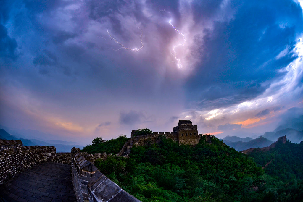 Great Wall of China under a dramatic sky with lightning in the background. Lush greenery surrounds the ancient structure creating a striking contrast