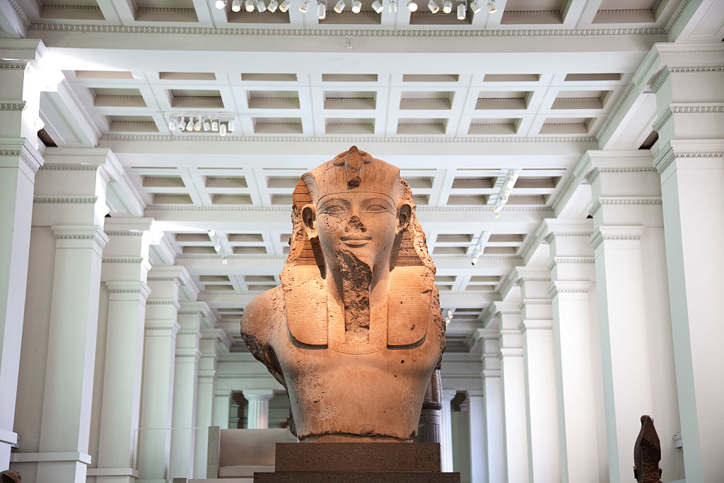 A large statue of an ancient Egyptian pharaoh's head and upper torso is displayed in a grand hall with a high ceiling and numerous columns