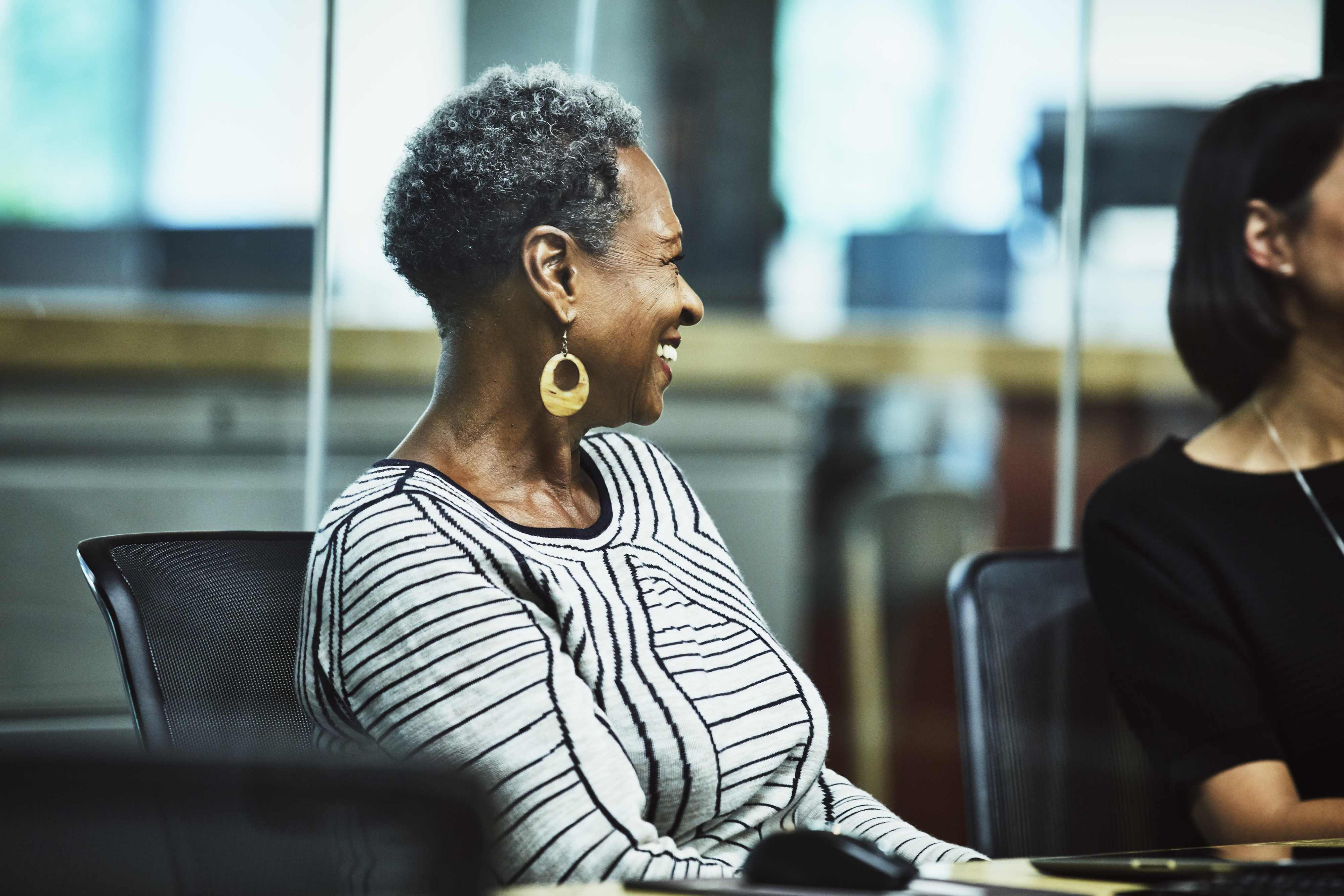 An older woman with short curly hair, wearing a striped sweater and large hoop earrings, smiles while seated in an office setting. Another person is partially visible