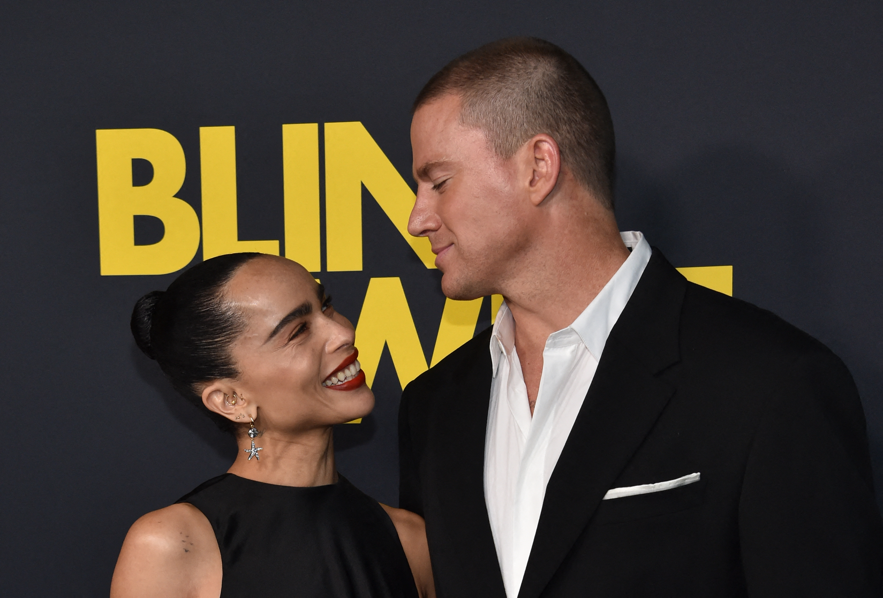 Zoë Kravitz and Channing Tatum smiling at each other at a movie premiere, Zoë in a sleeveless dress and Channing in a black suit with a white shirt