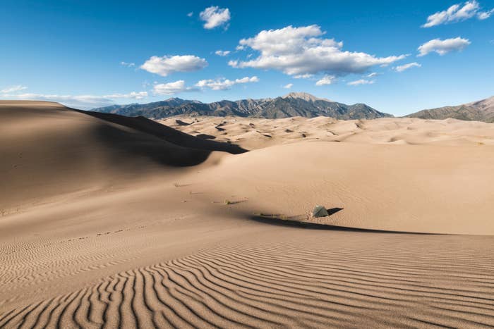 Vast sand dunes under a partly cloudy sky with mountains in the background. No people are visible