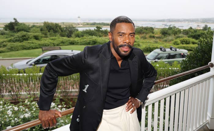 Colman Domingo stands on a porch with a scenic view in the background, dressed in a stylish jacket and pants