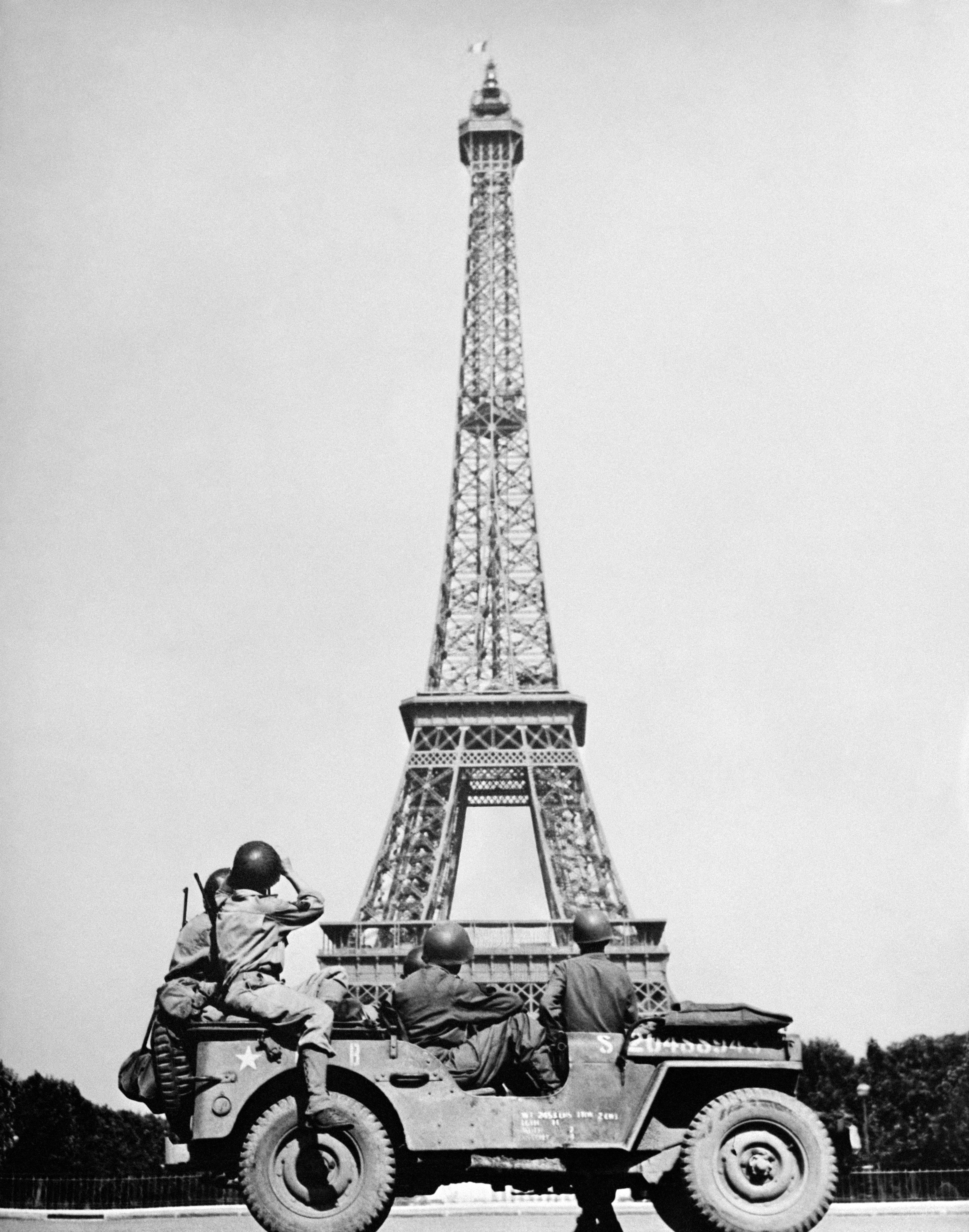 Three soldiers in a military jeep look at the Eiffel Tower in Paris, France from the base. The scene appears historical