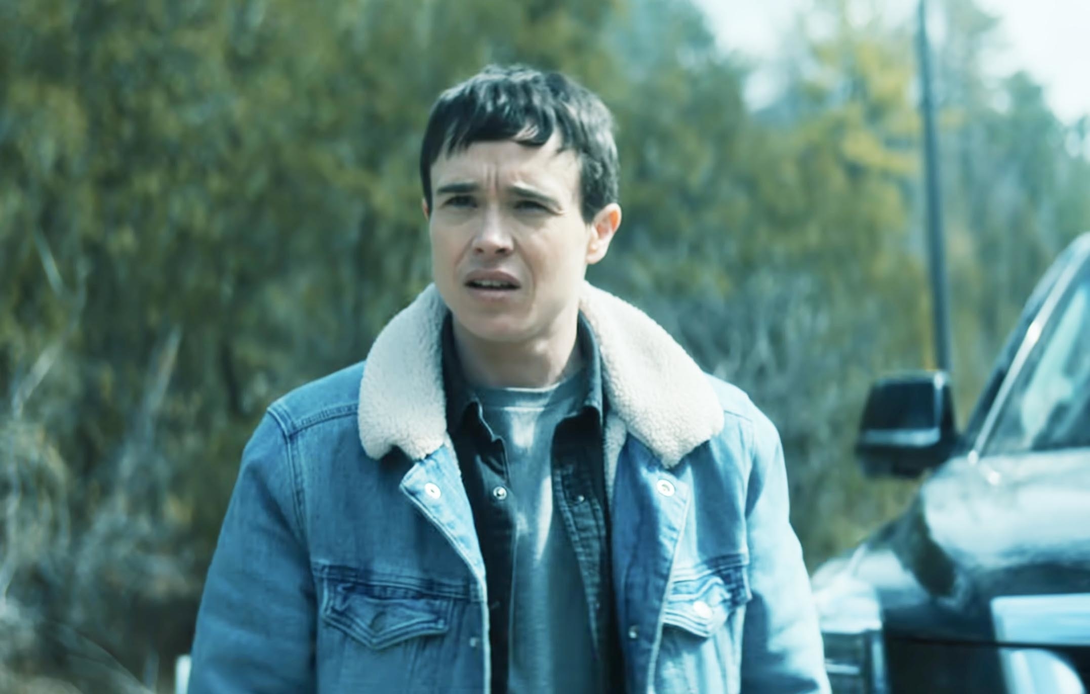 Elliot Page, dressed in a denim jacket with a shearling collar, stands outdoors with a concerned expression next to a vehicle