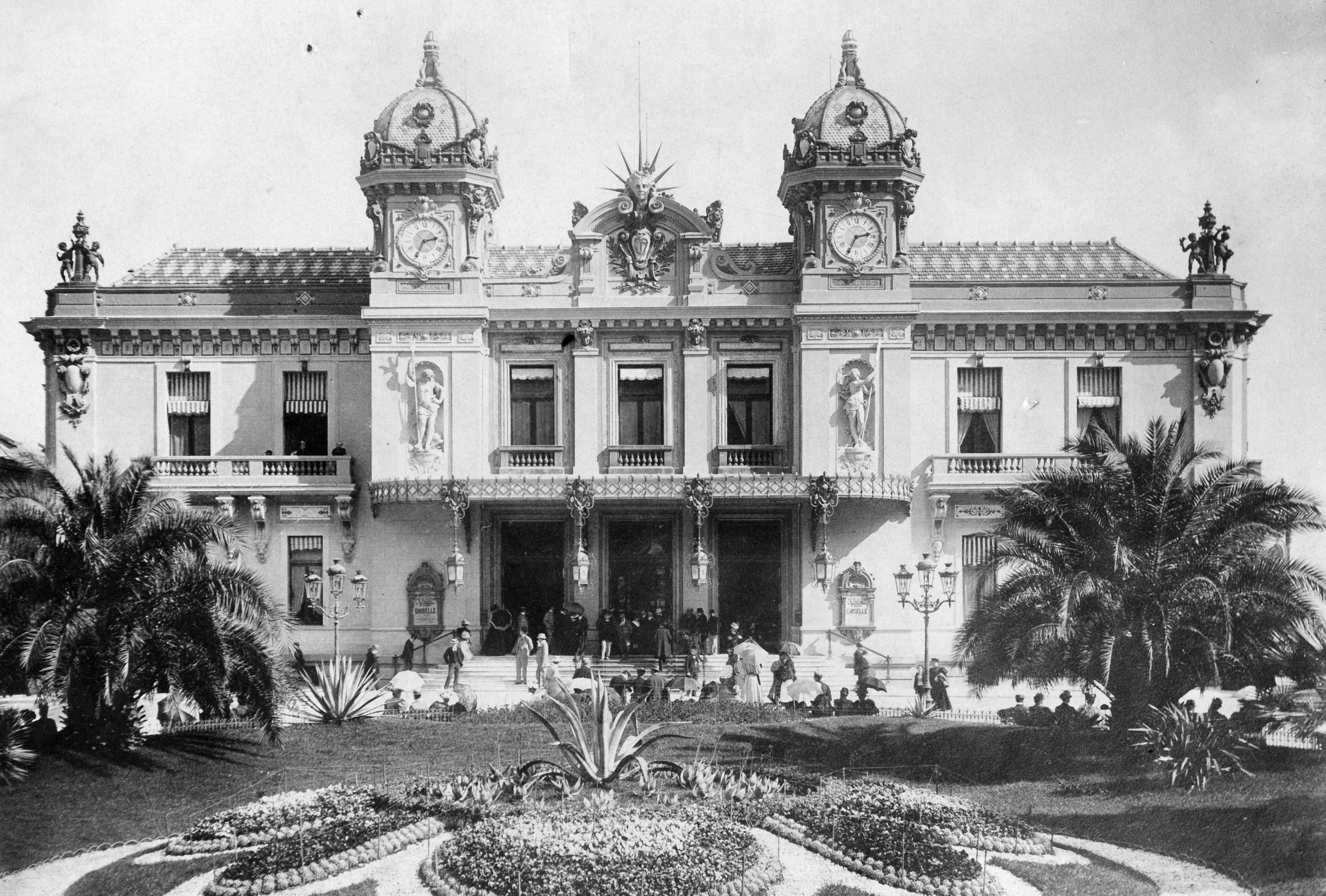 A historical black-and-white photograph of the Casino de Monte-Carlo, featuring people in front of the grand building with intricate architectural details and clock towers