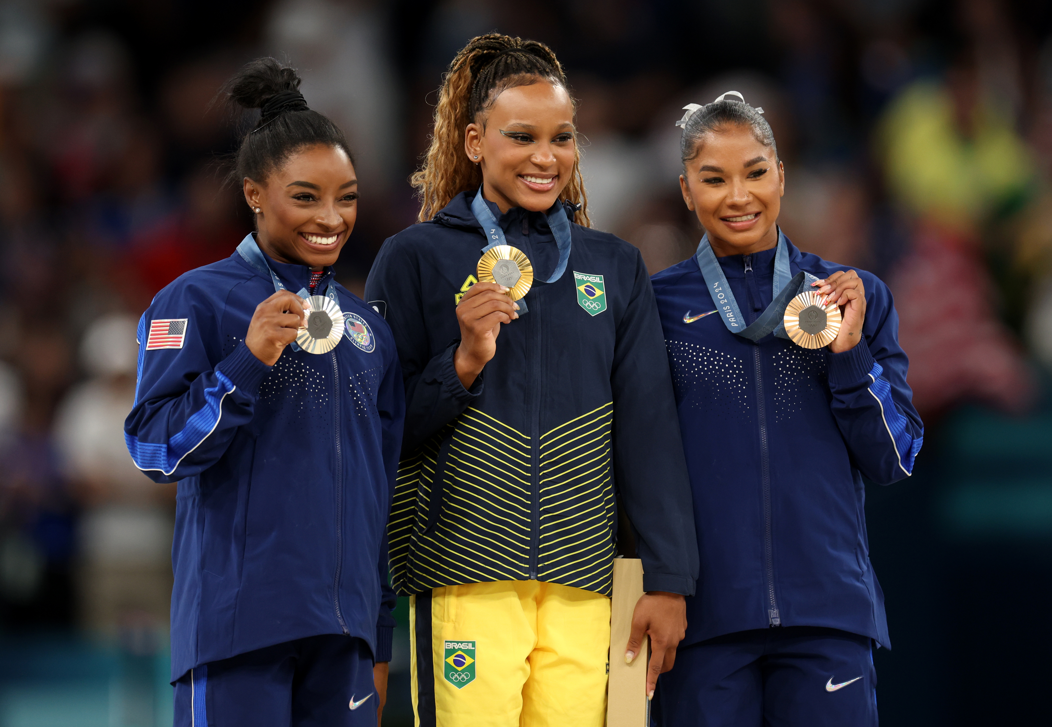 Simone Biles, Rebeca Andrade, and Sunisa Lee smiling and holding their medals on the podium at a gymnastics competition