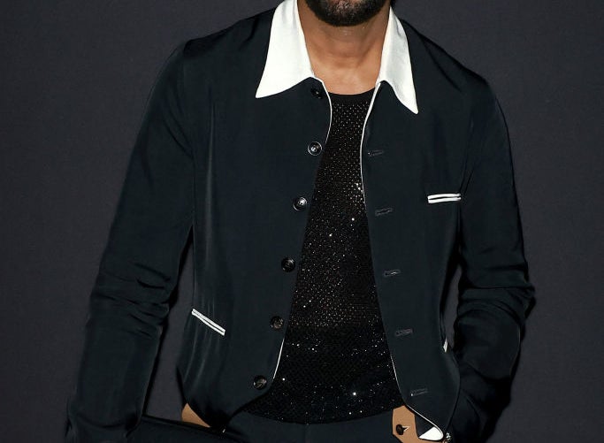 Colman Domingo smiles while posing in a stylish black outfit with a white collar and holding a black clutch