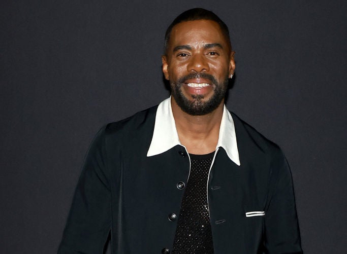 Colman Domingo smiles while posing in a stylish black outfit with a white collar and holding a black clutch
