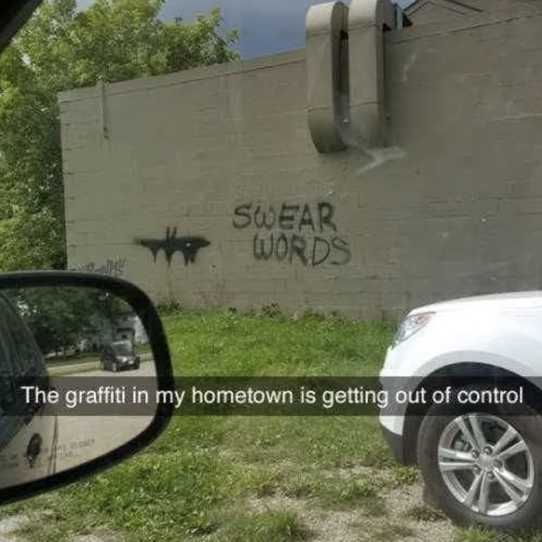 Graffiti on a wall reads "SWEAR WORDS" next to a Batman symbol. A vehicle's side mirror and part of a white SUV are visible. Text overlay says graffiti is out of control